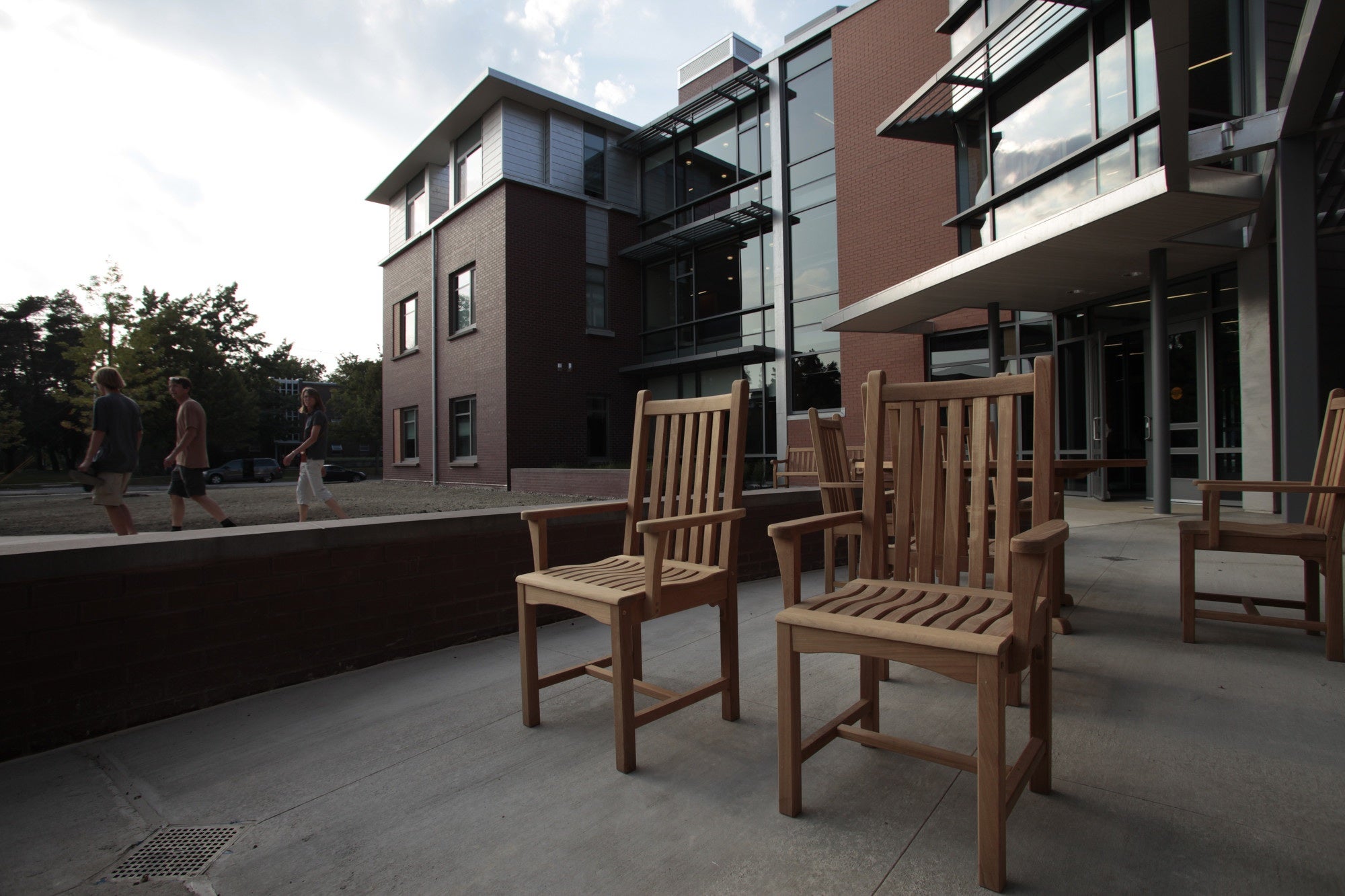 Wood chairs on a concrete patio outside a brick and glass building.